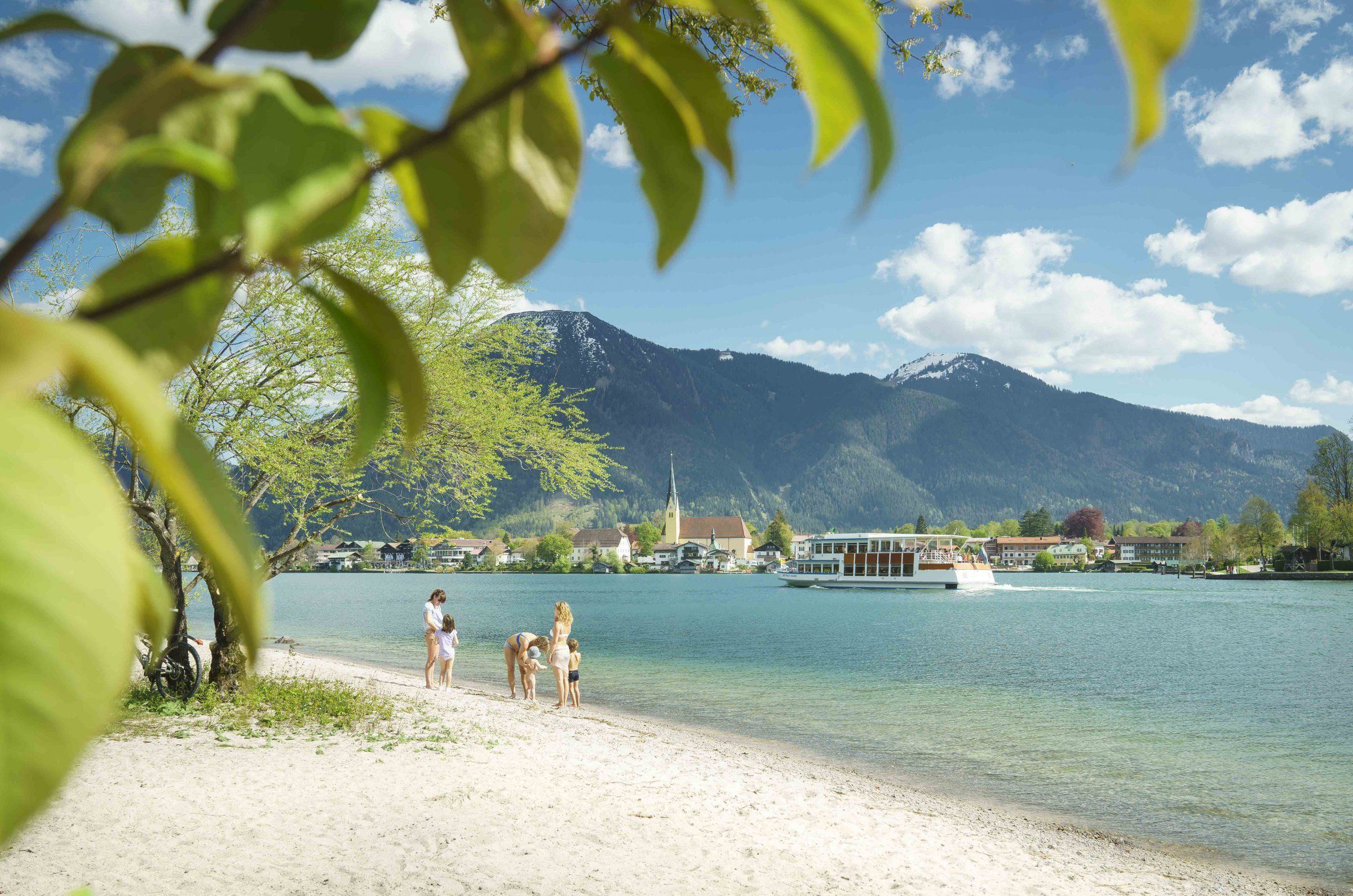 Ufer des Tegernsees mit Badegästen, einem Schiff und den Bergen dahinter