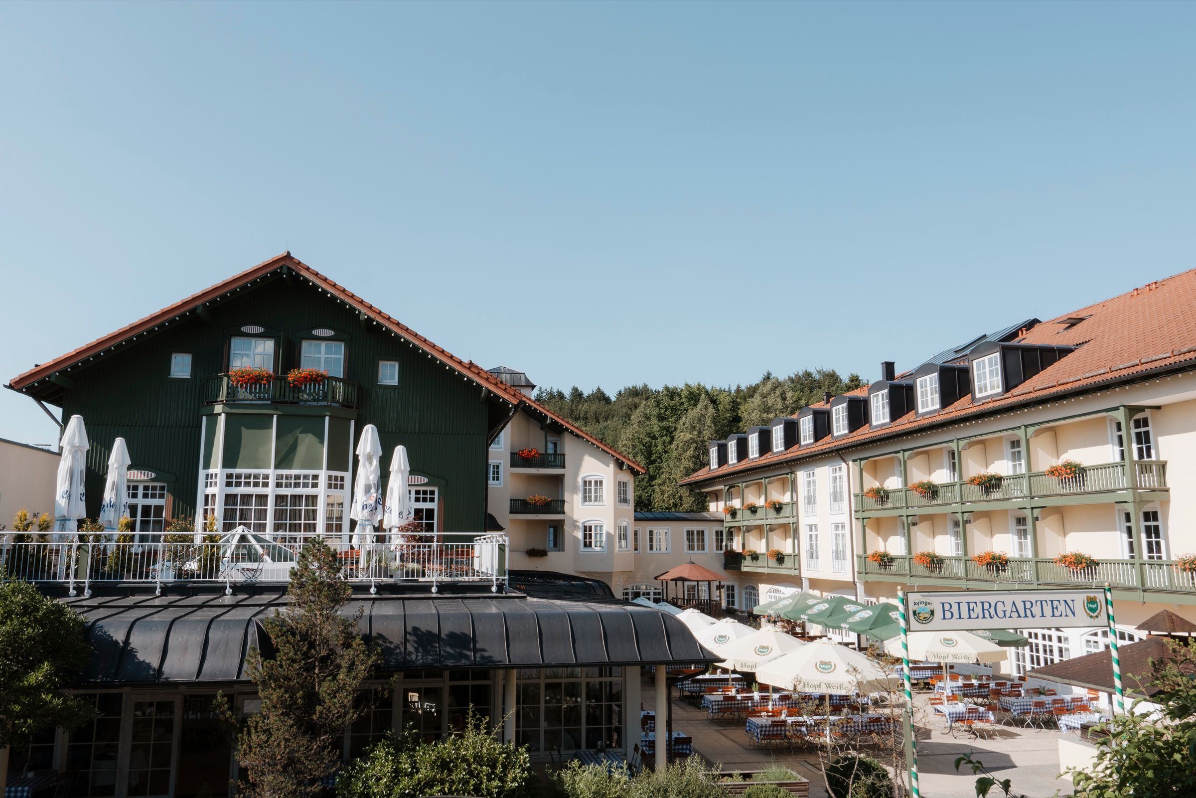 Der Bayerische Hof Miesbach: das grüne Haupthaus mit Holzbalkonen, Biergarten und Sonnenterrasse unter blauem Himmel