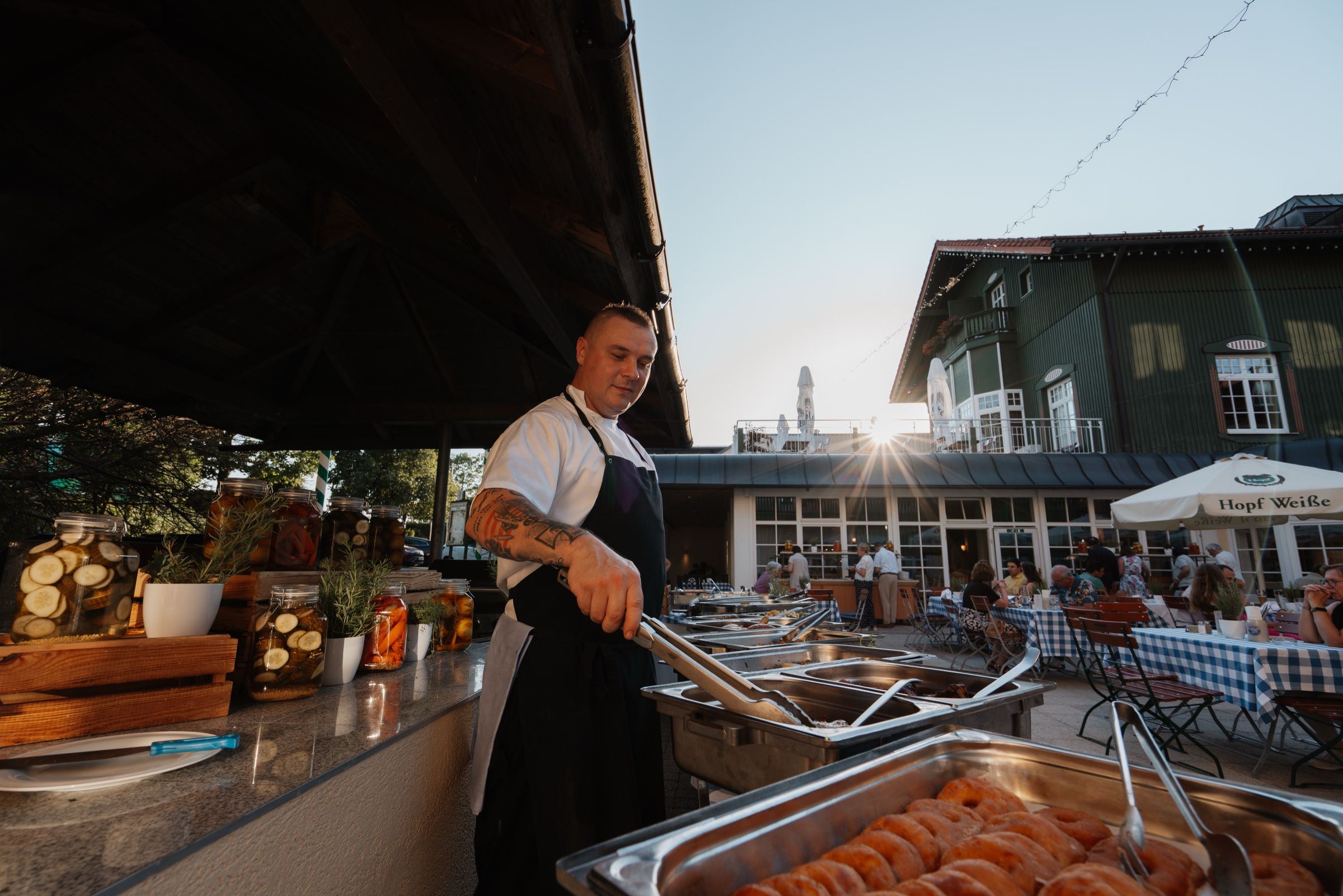Koch am Grill, dahinter Gäste im Biergarten in der Abendsonne